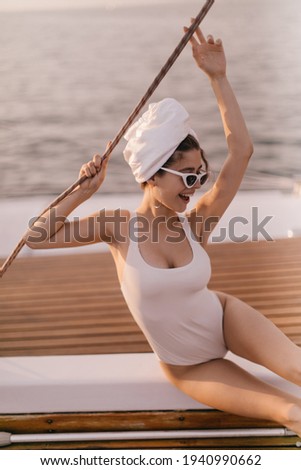 Excited young lady in white swimsuit, towel wrapped hair, stylish sunglasses and earrings, sitting on yacht deck and smiling against background of sea during sunset