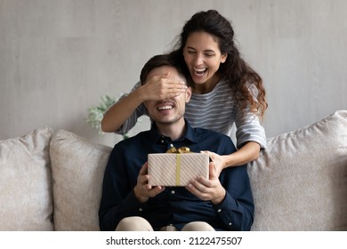 Excited young Latin woman giving surprise gift to excited boyfriend, standing aback, putting hand on eyes, laughing. Joyful millennial couple celebrating birthday, valentines day, anniversary