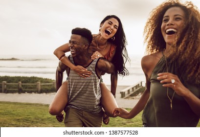 Excited young man carrying his girlfriend on his back with female friend walking in front. Diverse group of friends enjoying on a vacation. - Powered by Shutterstock