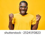 Excited young man celebrating success with a joyful expression, wearing stylish glasses and a yellow t shirt against a bright yellow background, full of vibrant energy and happiness