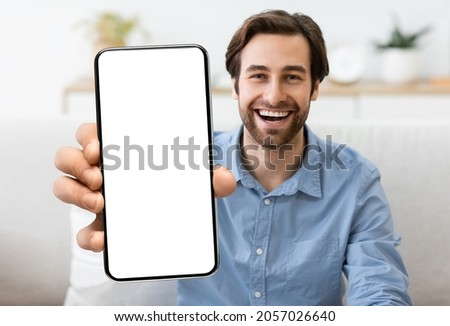 Excited Young Man Demonstrating Smartphone With Big Blank White Screen At Camera, Handsome Smiling Millennial Guy Showing Copy Space For Mobile App Advertisement, Sitting On Couch At Home, Mockup