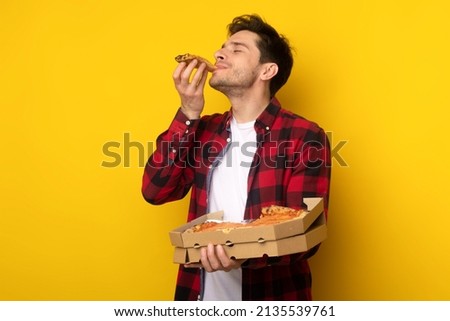 Excited Young Man Enjoying Pizza Holding And Biting Tasty Slice Posing Holding Carton Box On Yellow Orange Studio Wall. Junk Food Lover Eating Yummy Italian Pizza. Unhealthy Nutrition Cheat Meal