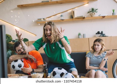 Excited Young Man In Green Fan T-shirt Throwing Popcorn And Celebrating While His Upset Friends Sitting And Gesturing Behind On Sofa During Watch Of Soccer Match At Home