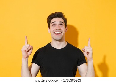 Excited Young Man Guy 20s In Casual Black T-shirt Posing Isolated On Yellow Wall Background Studio Portrait. People Sincere Emotions Lifestyle Concept. Mock Up Copy Space. Pointing Index Fingers Up