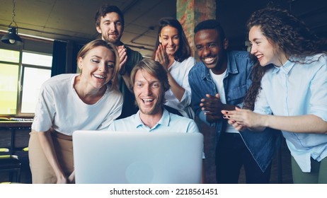 Excited Young Man Is Looking At Laptop Screen, Rejoicing And Expressing Happiness, His Happy Colleagues Are Congratulating Him On Success, Clapping Hands