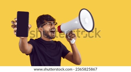 Excited young man on yellow studio background shouts in megaphone, advertises sale, recommends Internet store, promotes new model, draws attention to mobile phone with black mockup copy space screen
