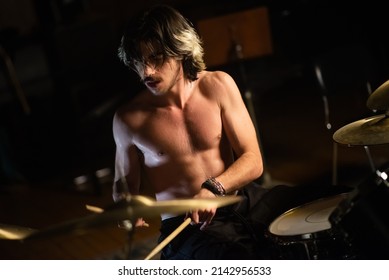 Excited Young Man Sitting Shirtless And Playing Drum Set In Garage. Rock Musician Training Or Recording Drums. Drummer Training Concept