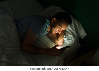 Excited Young Man Texting And Watching Online Videos On His Smartphone While Staying Up Late At Night