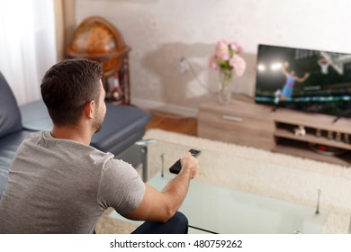 Excited Young Man Watching Basketball In Living Room, Pushing Button On Remote Control