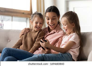 Excited Young Mixed Race Babysitter Cuddling Little Children, Looking At Smartphone Screen. Happy Small Girl Showing Funny Cartoon Photo On Mobile Phone To Laughing Mother And Sister, Resting At Home.