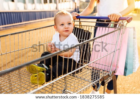 Excited young mother with baby and shopping bags and shopping trolley near mall