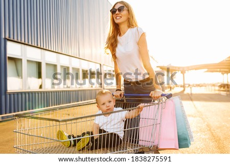 Excited young mother with a child and shopping bags fun running with a shopping trolley on the street near a shopping center