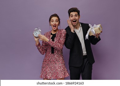 Excited Young Pair In Evening Party Outfits Emotionally Waiting For Holiday, Holding Alarm Clock And Gift Box, And Posing Isolated On Violet Background 