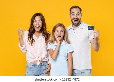 Excited Young Parents Mom Dad Child Kid Daughter Teen Girl In Basic T-shirts Hold Credit Bank Card Doing Winner Gesture Isolated On Yellow Background Studio Portrait. Family Day Parenthood Concept