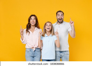 Excited Young Parents Mom Dad With Child Kid Daughter Teen Girl In Basic T-shirts Pointing Index Fingers Up On Mock Up Copy Space Isolated On Yellow Background Studio Portrait. Family Day Concept