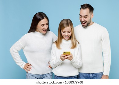 Excited Young Parents Mom Dad With Child Kid Daughter Teen Girl In White Sweaters Using Mobile Phone Typing Sms Message Isolated On Blue Background Studio. Family Day Parenthood Childhood Concept