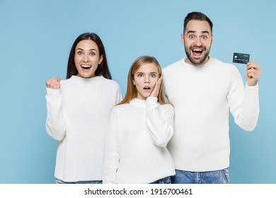 Excited Young Parents Mom Dad With Child Kid Daughter Teen Girl In White Sweaters Hold Credit Bank Card Doing Winner Gesture Isolated On Blue Background Studio Portrait. Family Day Parenthood Concept