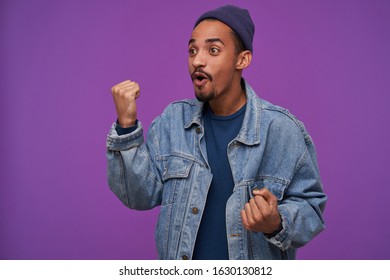 Excited young pretty dark haired bearded male showing fist while looking aside with agitated face, wearing blue cap, pullover and jeans coat while posing over purple background