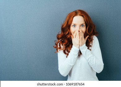 Excited young redhead woman staring wide eyed at the camera as she covers her mouth with her hands over a dark studio background with copy space