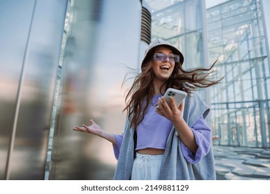 Excited Young Smiling Woman Holding And Using Mobile Phone. Cool Stylish Girl At Outside