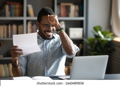 Excited Young Successful African American Man Feeling Overjoyed About Win Paper Notification, Sitting At Table With Laptop. Happy Mixed Race Businessman Celebrating Triumph, Reading Letter With News.