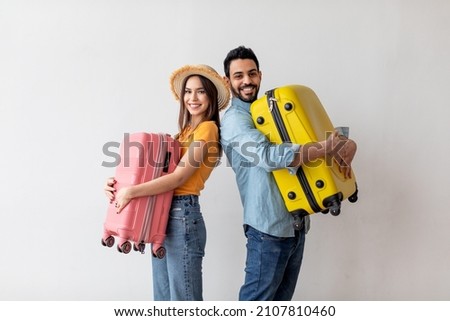 Excited young tourists couple ready for traveling abroad, holding suitcases in hands and smiling at camera, standing over light wall. Air flight journey concept