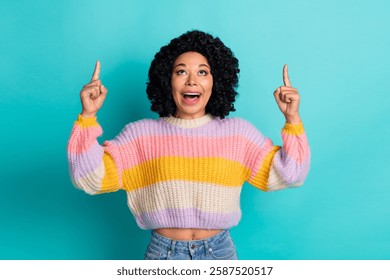 Mujer joven emocionada con el pelo rizado negro que lleva un suéter colorido que muestra los gestos que señalan en el fondo verde azulado