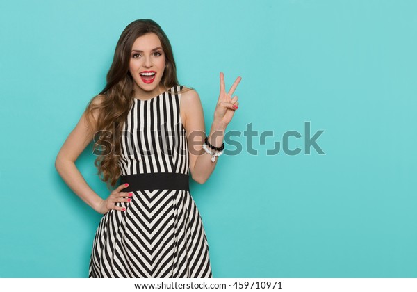 Excited young woman in black and white\
striped dress showing peace hand sing. Three quarter length studio\
shot on teal\
background.