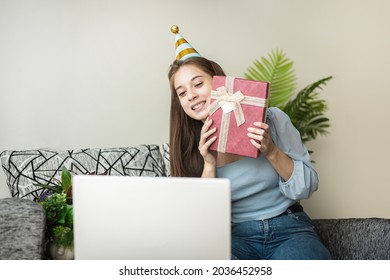 Excited Young Woman Holding And Opening Christmas Present, Unwrapping Gift Box , Sitting On The Sofa At Home In Living Room With Laptop Computer, Celebrating Online