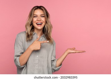 Excited young woman holding palm open for new product or sale presentation. Overjoyed happy blond girl with open mouth point finger at arm with copy space for promotion isolated over pink studio wall - Powered by Shutterstock