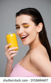 excited young woman holding yellow can with soft drink and winking eye isolated on grey