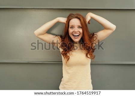Excited young woman laughing at the camera with hands raised to her head against a grey wall