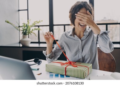 Excited Young Woman Office Employee Closing Eyes With Hand Unpacking Xmas Present Pulling Ribbon When Sitting At Workplace, Happy Freelancer Lady Opening Gift Box With Excitement During Remote Work