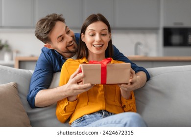 Excited young woman receiving surprise gift from man standing from back lady sitting on sofa,both smiling and capturing moment of joy and affection - Powered by Shutterstock