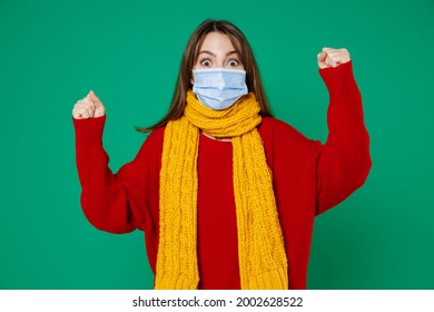 Excited young woman in red sweater yellow scarf sterile face mask to safe from coronavirus virus covid-19 during pandemic quarantine doing winner gesture isolated on green background studio portrait