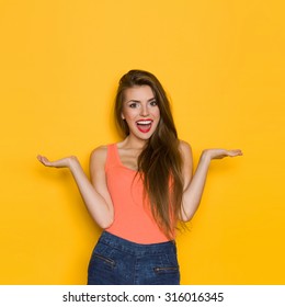 Excited Young Woman. Shouting young woman in orange shirt and jeans shorts posing with arms outstretched. Three quarter length studio shot on yellow background.