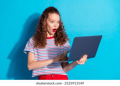 Excited young woman in striped shirt surprised while looking at laptop against blue background - Powered by Shutterstock