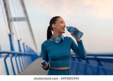 Excited young woman taking break from jogging, having break and drinking water, holding bottle to refresh herself during outdoor workout in the evening - Powered by Shutterstock