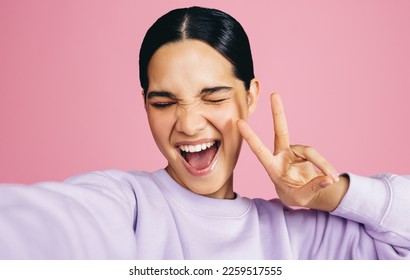 Excited young woman taking a selfie in a studio, she makes a peace sign with an expression of joy on her face. Woman having fun capturing her moments of self confidence and positivity. - Shutterstock ID 2259517555