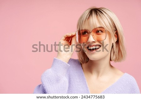 Excited young woman wearing sunglasses and braces, looking away. Positive female student posing isolated on pink background. Summer concept, advertisement 