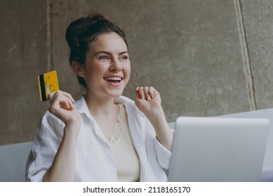 Excited Young Woman In White Clothes Sit On Grey Sofa Indoors Apartment Use Mobile Cell Phone Credit Bank Card Shopping Online Order Delivery Booking Tour Rest On Weekend Leisure Stay Home Concept.