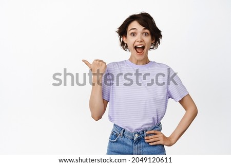 Excited young woman wow face, pointing thumb left and looking amazed, hey check this out, big announcement, showing discount sale, company logo, standing over white background