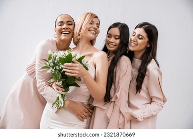 excitement, multicultural bridesmaids hugging happy bride in wedding dress with bridal bouquet, grey background, racial diversity, fashion, brunette and blonde, group of women, white flowers - Powered by Shutterstock