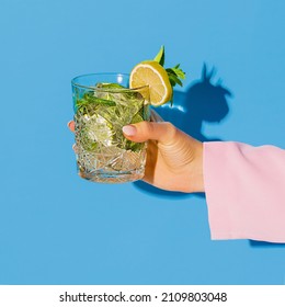 Exclusive Alcoholic Drink. Female Hand Holding Glass With Cocktail Mojito With Lemon Isolated On Light Blue Neon Background. Complementary Colors, Blue, Yellow And Green. Copy Space For Ad, Design