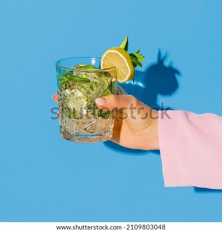 Exclusive alcoholic drink. Female hand holding glass with cocktail mojito with lemon isolated on light blue neon background. Complementary colors, blue, yellow and green. Copy space for ad, design