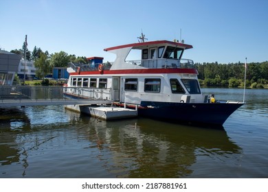 Excursion Tourist Steamer At The Pier. Nature, Dam. White Excursion Steamer With A Submersible Part In Blue. The Floor Of The Steamer Is Divided By A Red Stripe. Summer, Travel, Experiences.