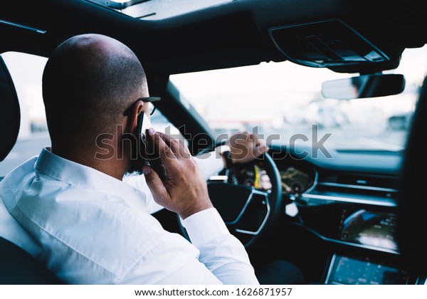 Executive businessman using international\
roaming connection for making cellphone call during wheel driving\
in luxury rent car, male communicating with partners while steering\
vehicle automobile