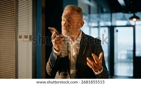 Executive dictating audio notes on smartphone walking corridor lit by sunset light closeup. Elderly businessman recording voice cellphone message in office hallway. Bearded manager talking loudspeaker