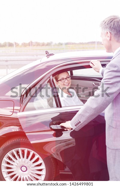 Executive holding car door for smiling colleague\
against sky