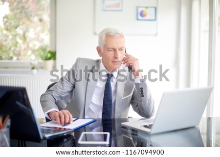 Executive senior businessman using his mobile phone and talking wih somebody while working laptop in the office. 